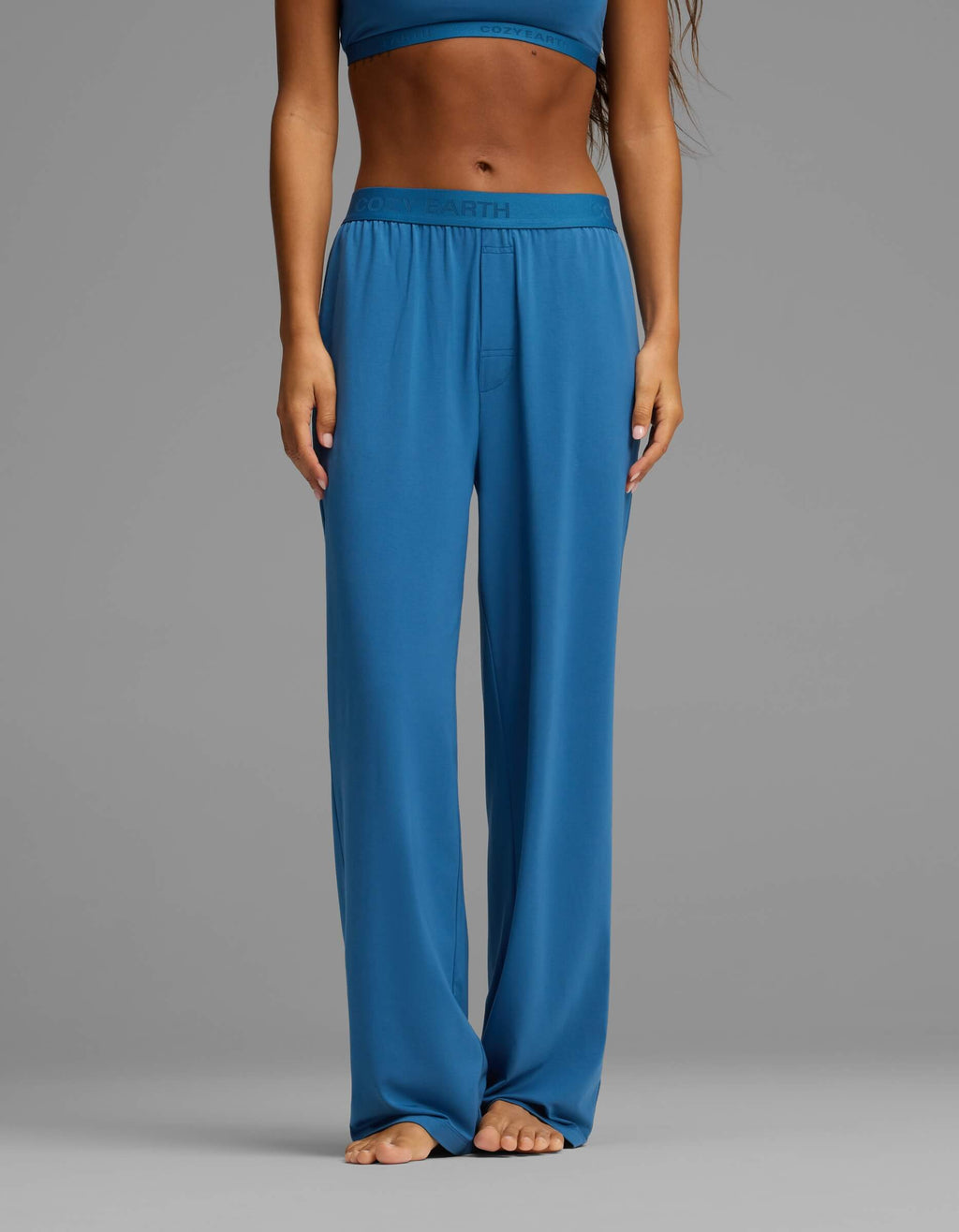 A person stands barefoot, wearing Cozy Earth’s Women’s First Layer Pajama Pant in blue with a matching top. The image is cropped from the shoulders down, with hands resting at the sides against a plain gray background.|Color:Anchor
