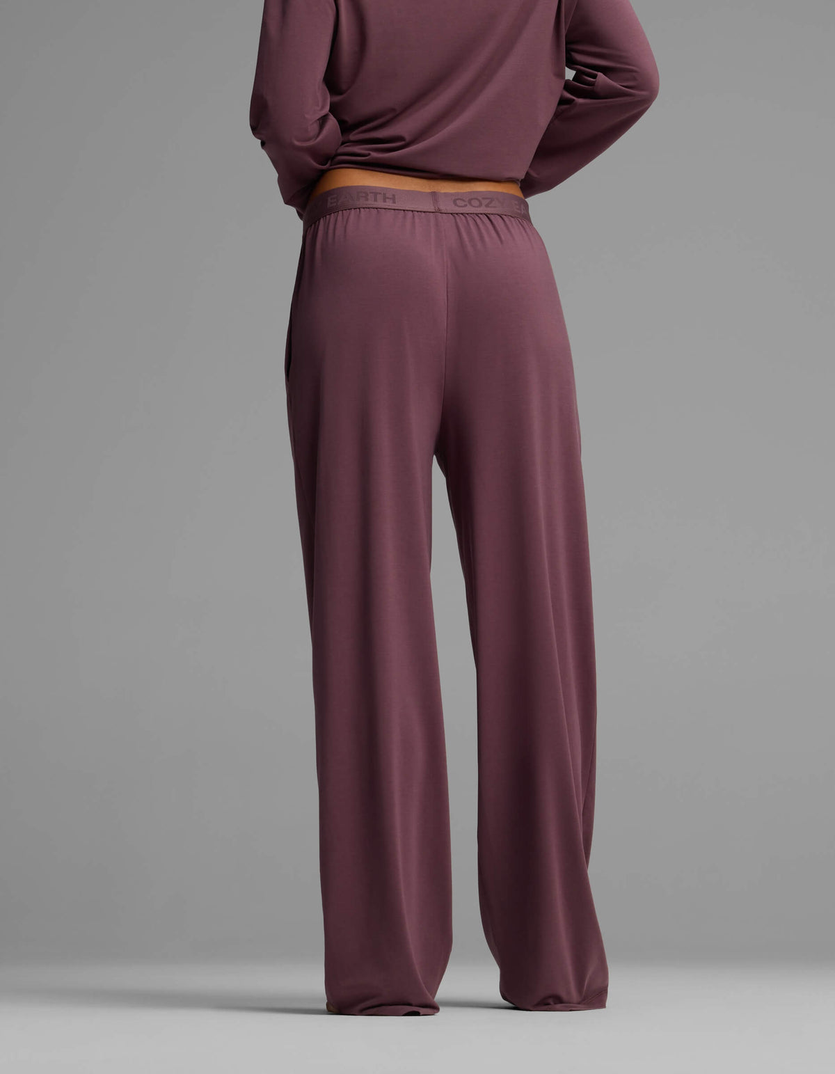 A woman is seen from the back wearing Cozy Earth’s Women’s First Layer Pajama Pant in a loose mauve fit, paired with a matching long-sleeve top, set against a plain gray background.|Color:Fig
