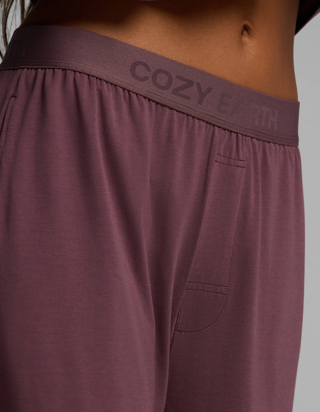 Close-up of a person wearing the Women’s First Layer Pajama Pant from Cozy Earth, featuring “COZY EARTH” printed on the waistband and a visible side pocket.|Color:Fig
