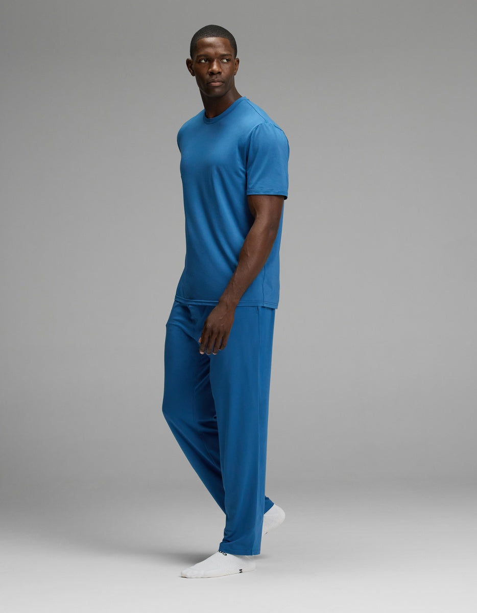 A man stands in a relaxed pose wearing the Cozy Earth Men’s First Layer Pajama Short Sleeve Tee & Pant Set in blue, paired with white shoes, against a plain gray background.|Color:Anchor