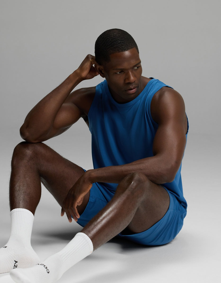 A man models the Cozy Earth Men’s First Layer Pajama Tank & Short Set in blue, sitting on the floor with one hand resting on his head and looking to the side. He completes the look with white socks against a plain, light gray background.|Color:Anchor