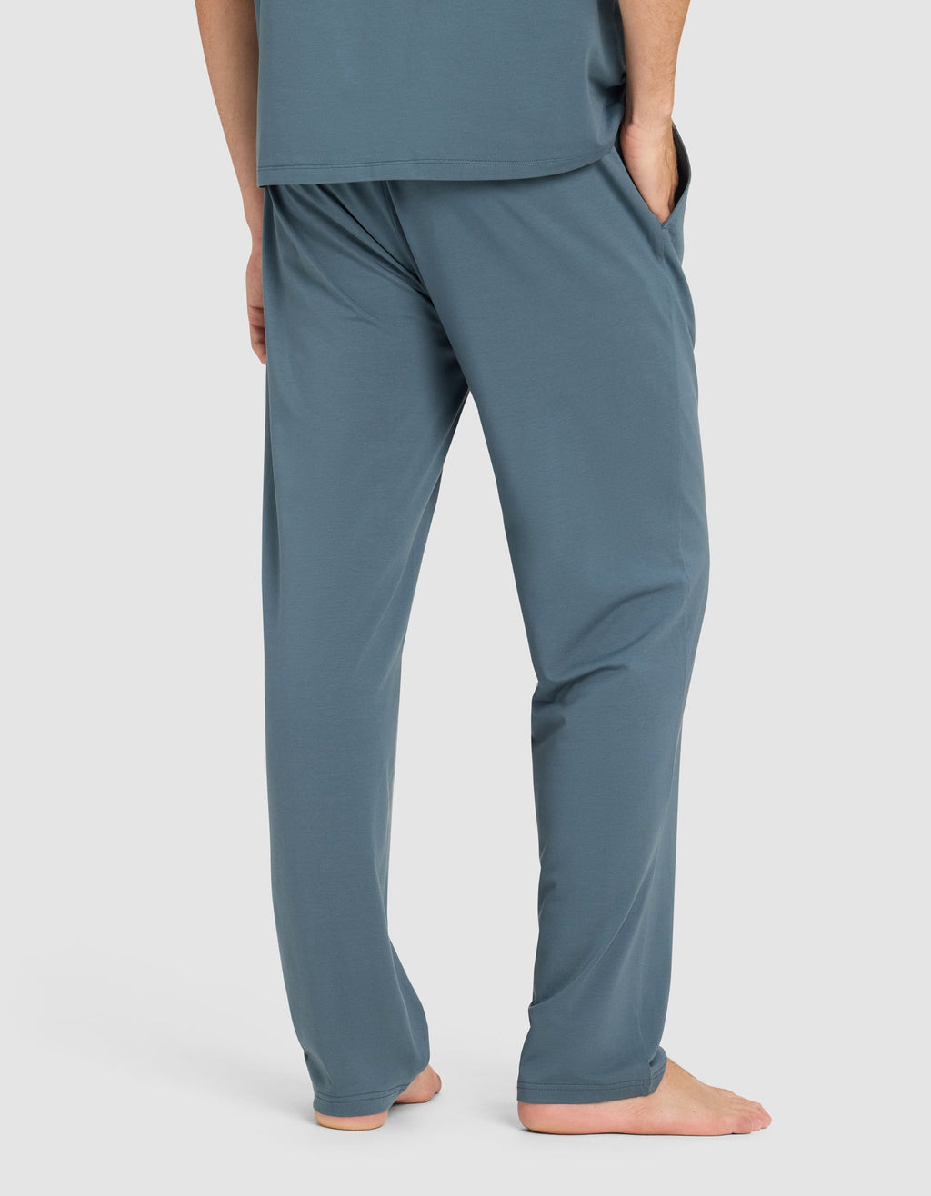 A man stands barefoot with hands in pockets, wearing Cozy Earth's Men's First Layer Pajama Pant in teal blue, paired with a matching top. He is shown from the waist down against a plain light background.|Color:Dawn