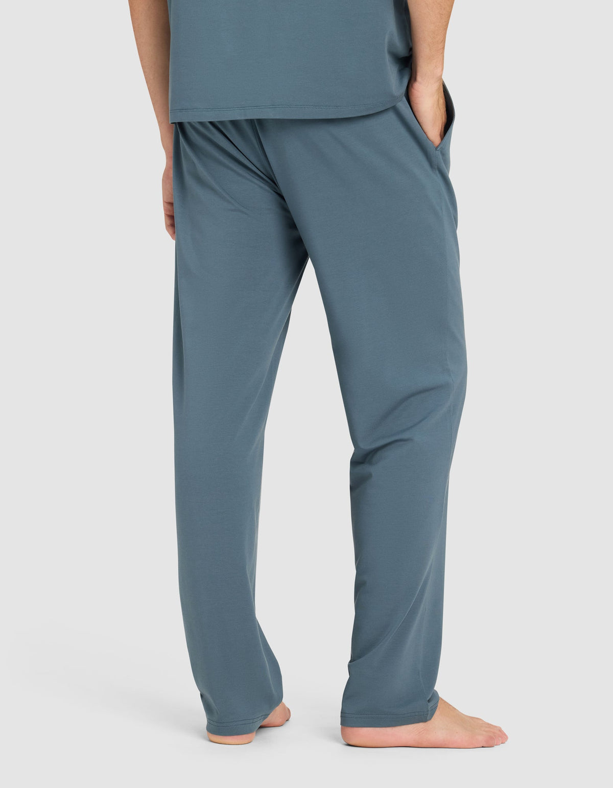 A man stands barefoot with hands in pockets, wearing Cozy Earth's Men's First Layer Pajama Pant in teal blue, paired with a matching top. He is shown from the waist down against a plain light background.|Color:Dawn