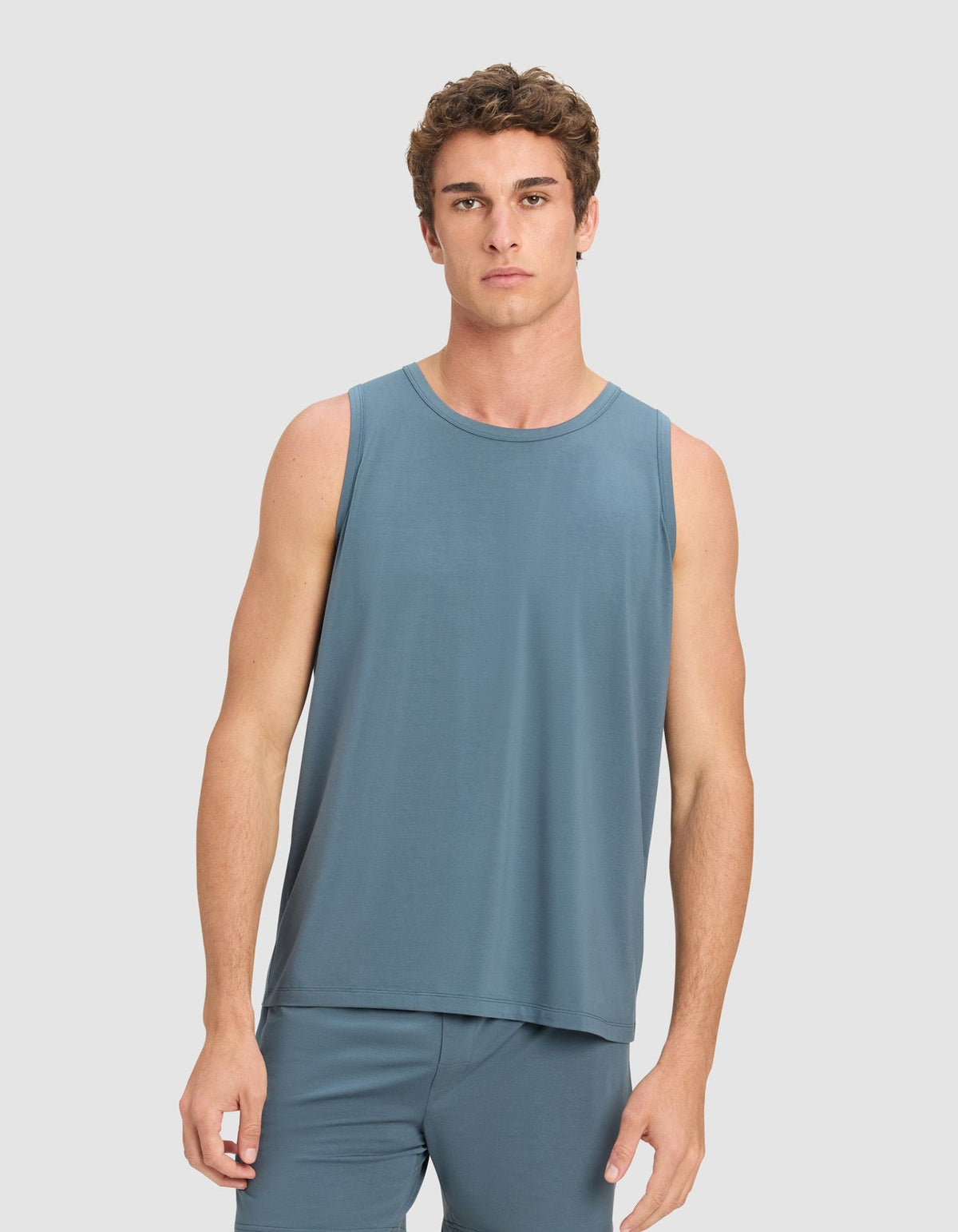 A young man with short, curly brown hair wears the Cozy Earth Men's First Layer Pajama Tank in blue, paired with matching shorts, standing against a plain light gray background and looking directly at the camera.|Color:Dawn