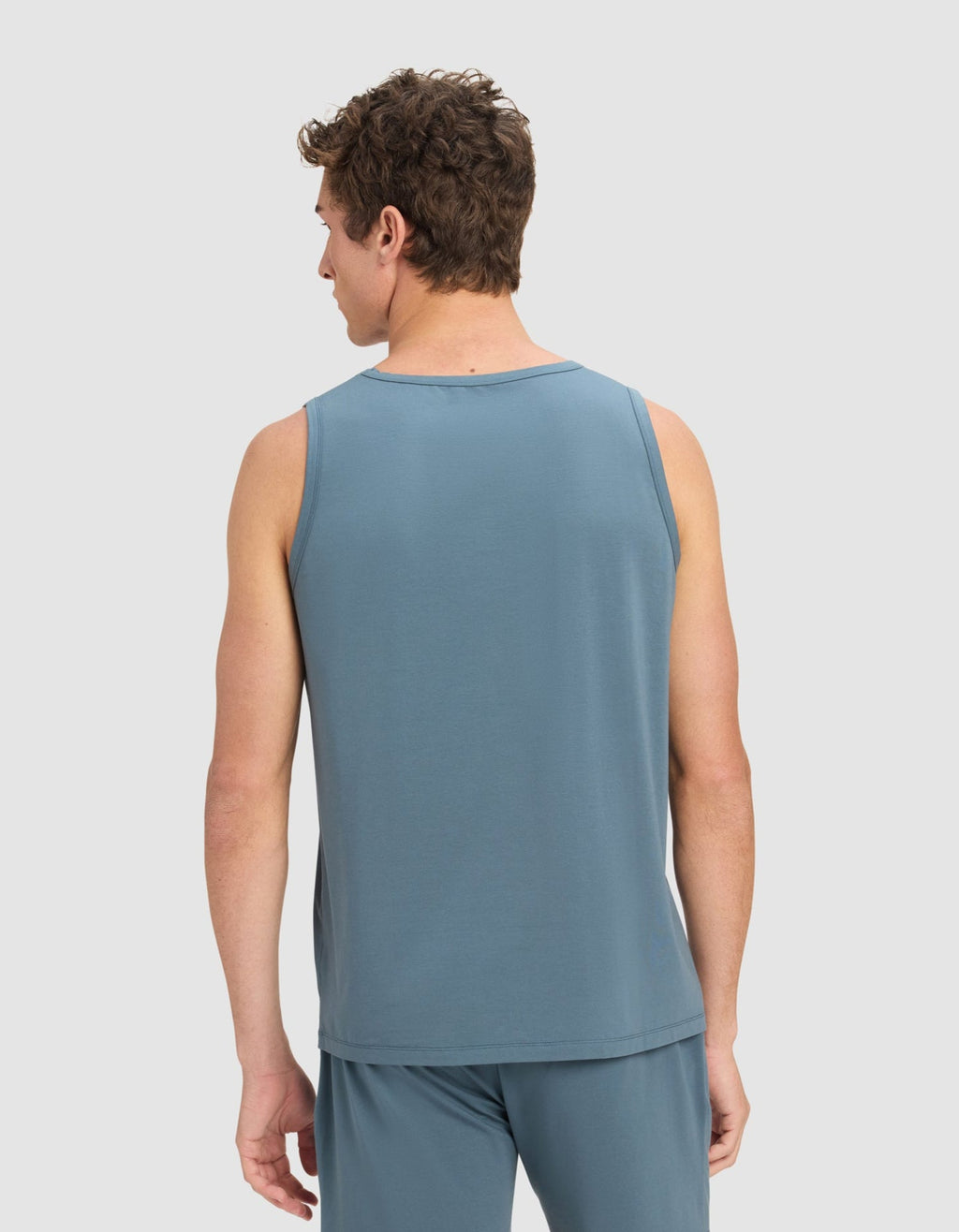A man faces away from the camera wearing Cozy Earth’s Men's First Layer Pajama Tank in blue, paired with matching shorts. The background is plain and light-colored.|Color:Dawn