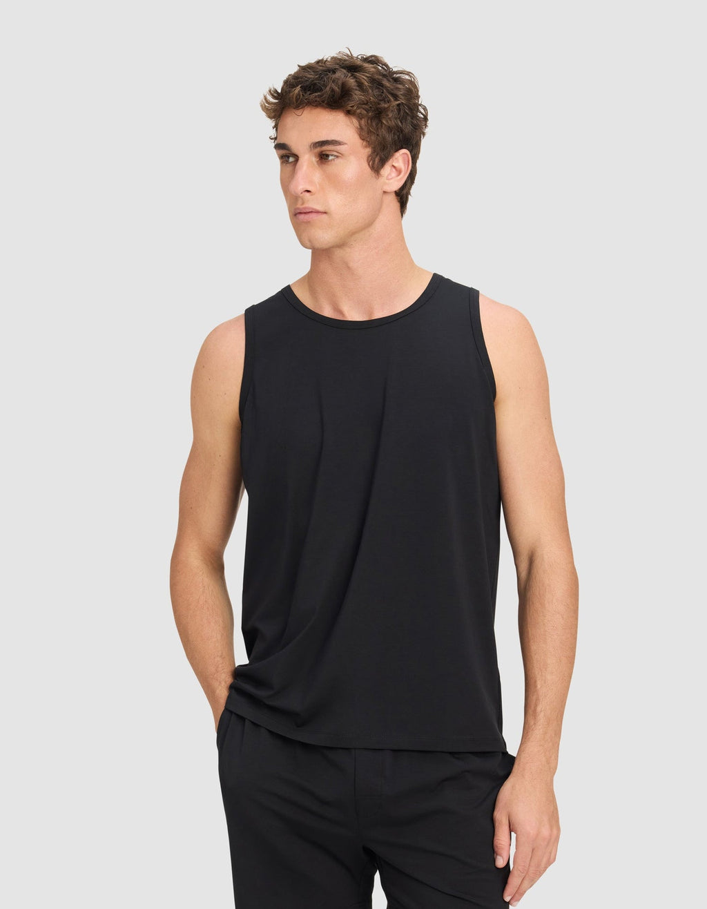 A man wears the Men's First Layer Pajama Tank by Cozy Earth and black shorts, standing with one hand in his pocket against a plain light gray background.|Color:Jet Black