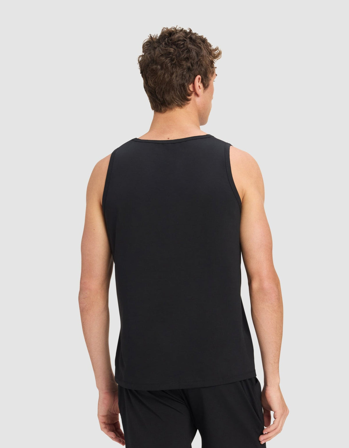 A man with short curly hair is seen from the back wearing Cozy Earth's Men's First Layer Pajama Tank in black, paired with black shorts, standing against a plain light gray background.|Color:Jet Black