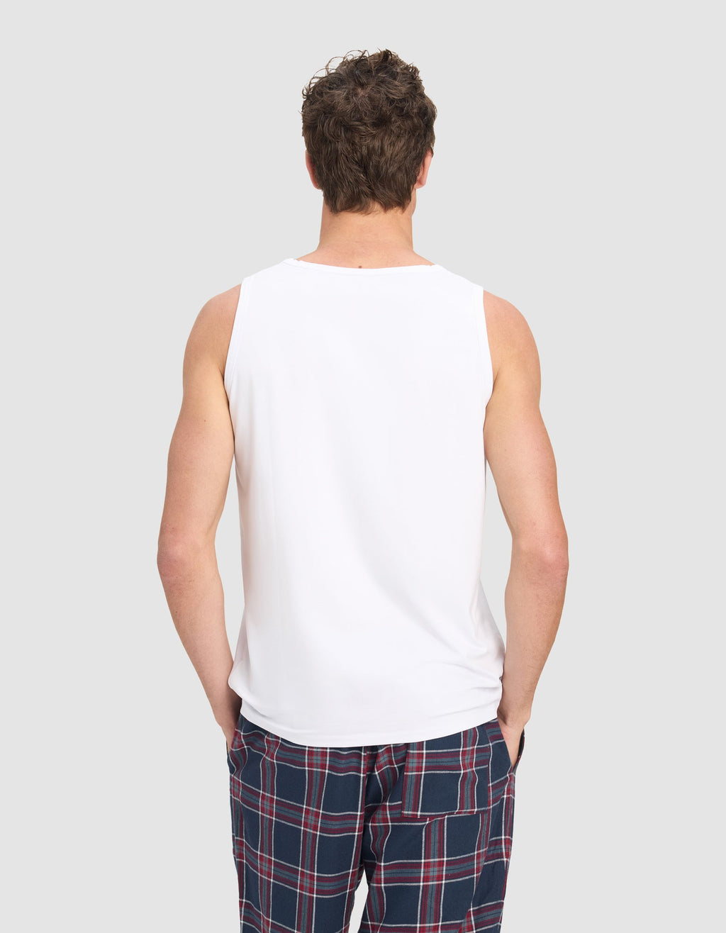 A man models the Cozy Earth Men's First Layer Pajama Tank in white with blue plaid pajama pants featuring red and white stripes, standing against a plain light gray background.|Color:White