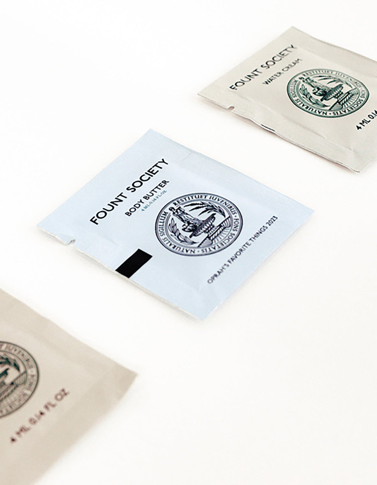 Three small, flat packets from the Cozy Earth Fount Society Intro Kit are arranged on a white surface. The nearest packet reads "BODY BUTTER" and each displays a circular emblem with product details in minimal black text.