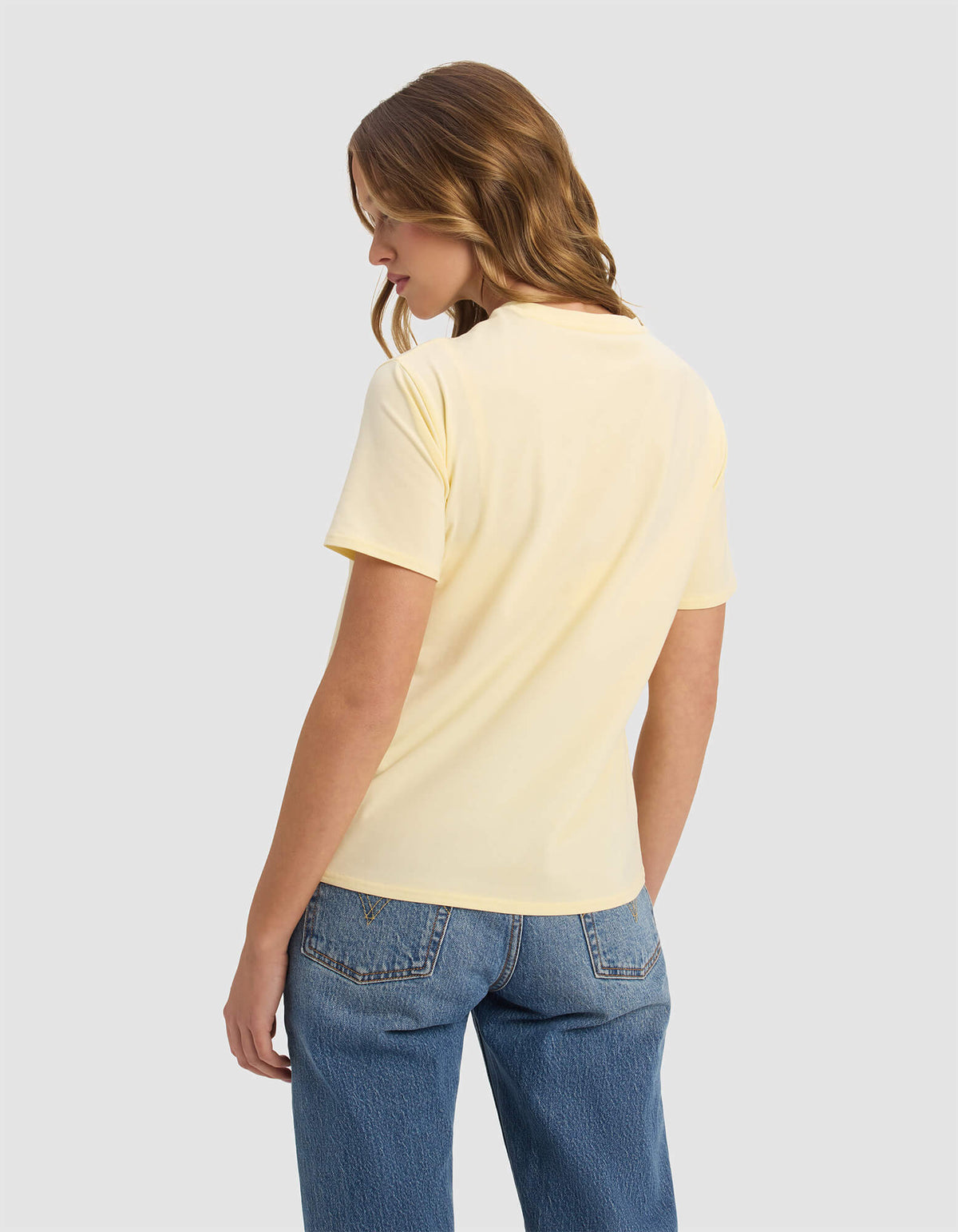 A woman with wavy light brown hair models the Cozy Earth Women's Go-To Tee in pale yellow, paired with blue jeans, as she stands facing away and glances over her left shoulder against a plain light gray background.|Color:Meringue