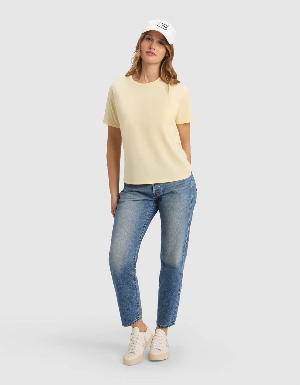 A woman stands with one hand in her pocket, wearing the Cozy Earth Women's Go-To Tee in pale yellow, a white cap, blue jeans, and white sneakers against a plain light gray background.|Color:Meringue