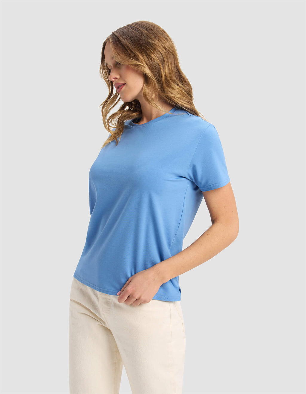 A woman with wavy light brown hair wears the Cozy Earth Women's Go-To Tee in light blue and off-white pants, standing against a plain light grey background with one hand in her pocket, looking down.|Color:Sailor