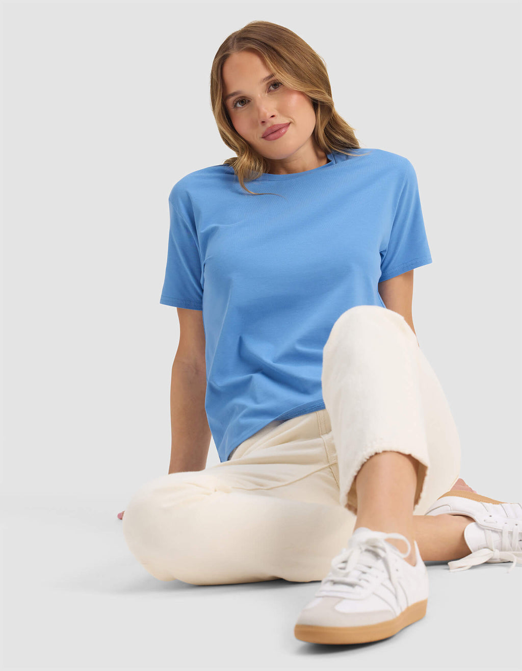 A woman with shoulder-length light brown hair sits against a plain background, looking relaxed with one knee raised. She wears Cozy Earth's Women's Go-To Tee in light blue, paired with cream pants and white sneakers, sporting a slight smile.|Color:Sailor