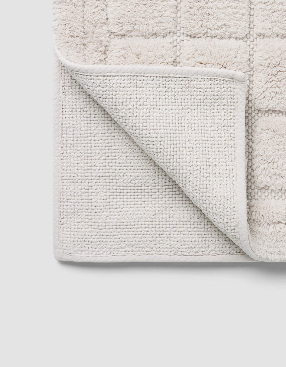 A close-up of the Cozy Earth Grid Bath Mat shows its light color, textured grid pattern, and one corner flipped to reveal the woven underside.|Color:Light Grey