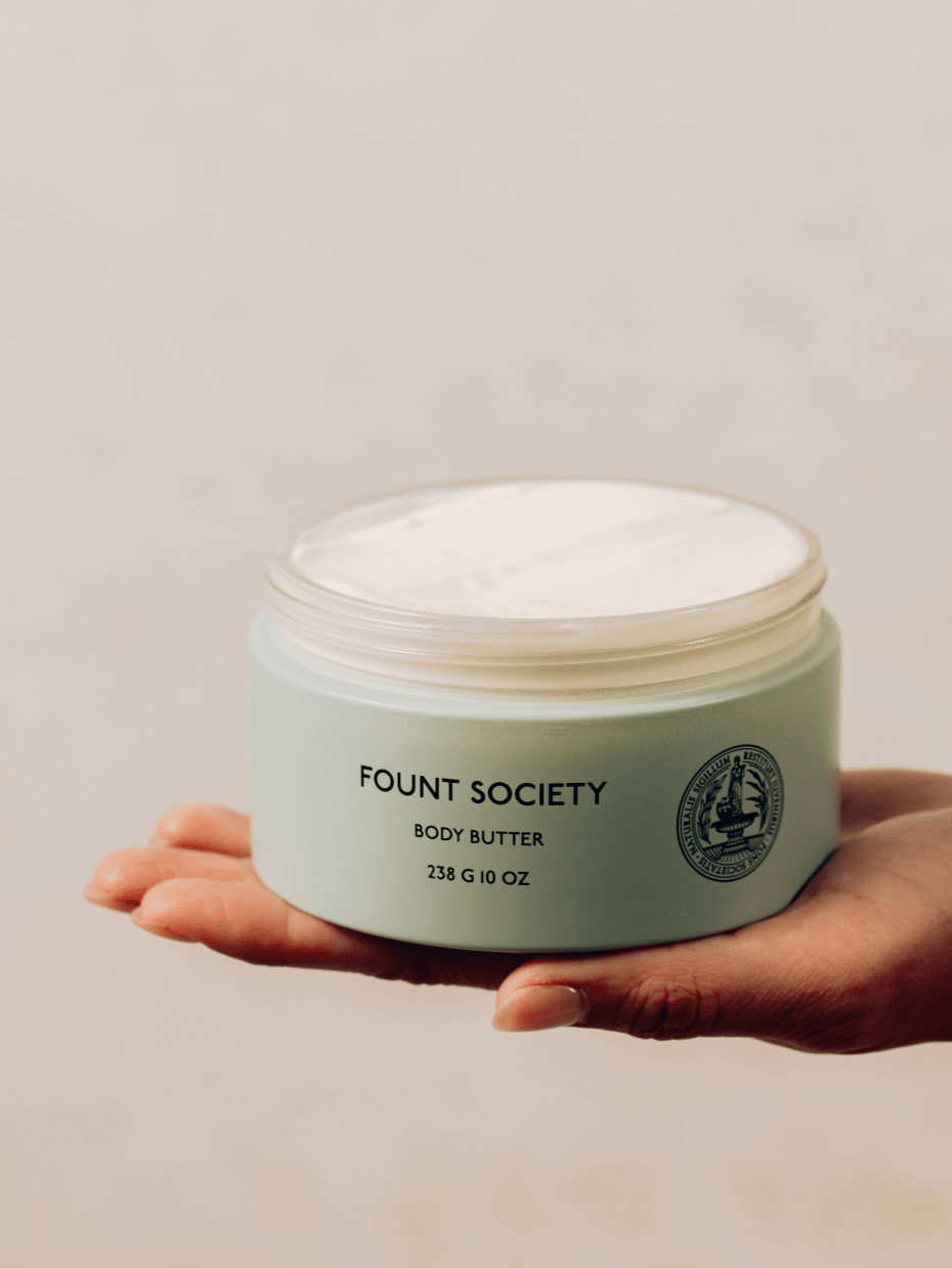 A hand holding a light green container of Cozy Earth Body Butter with a slightly opened lid, revealing the creamy product inside. The container has a 238 g (10 oz) label and a circular emblem. The background is softly blurred.|Size:Full Size