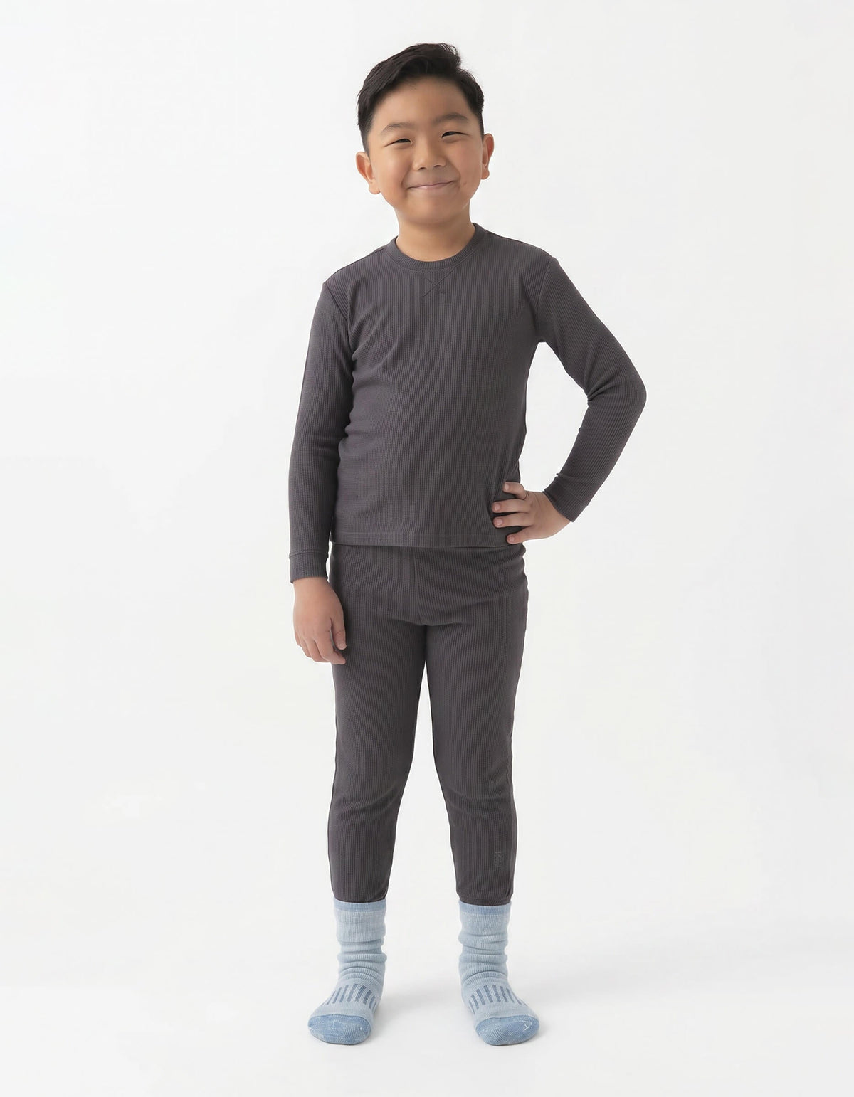 A young boy smiles with one hand on his hip, wearing the Cozy Earth Children's Bamboo Waffle Knit Base Layer Set in dark gray, with light blue socks, standing against a plain white background.