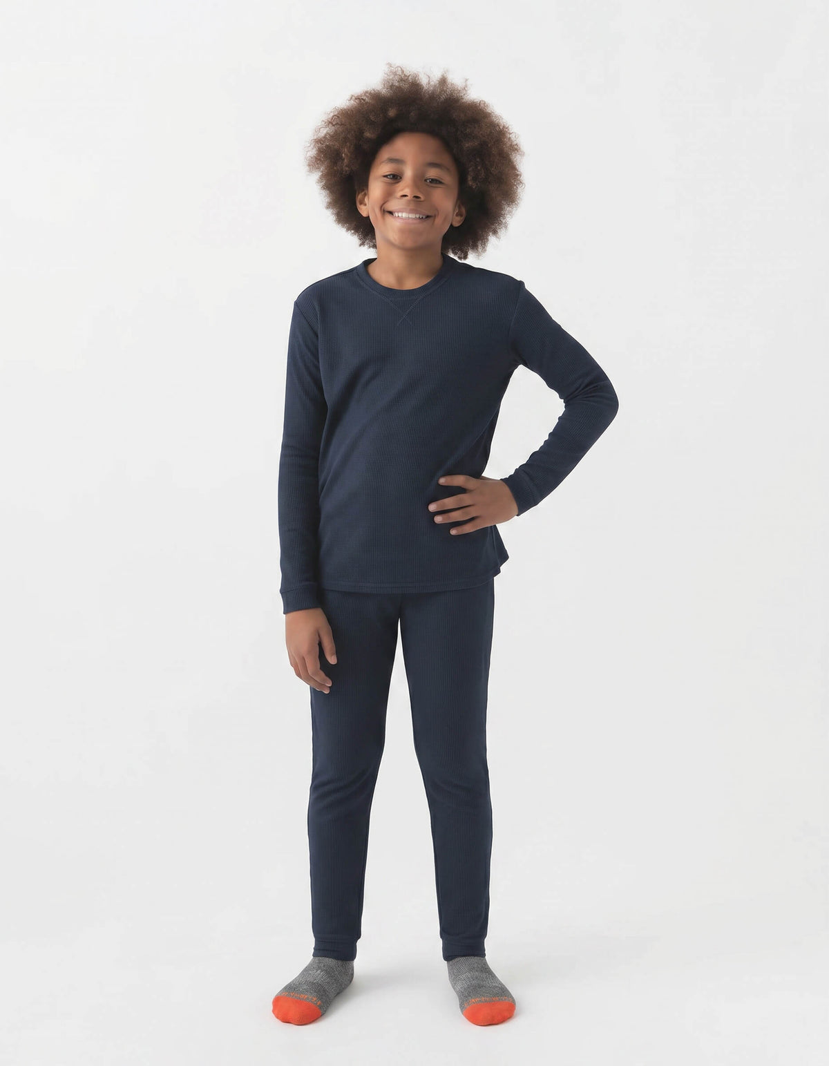 A smiling child with curly hair stands against a white background, wearing Cozy Earth's Children's Bamboo Waffle Knit Base Layer Set—navy top and pants—paired with gray and orange socks. One hand rests on their hip.|Color:Navy