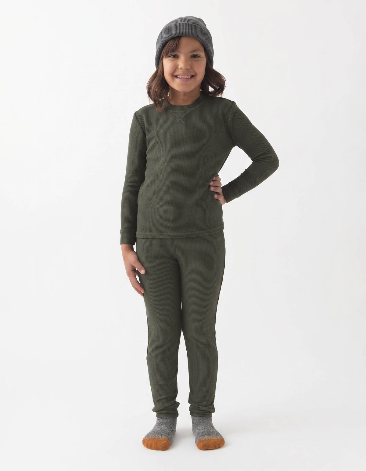 A young child smiles against a white background, wearing Cozy Earth's Children's Bamboo Waffle Knit Base Layer Set in dark green, paired with a gray beanie and gray socks with brown toes.|Color:Olive