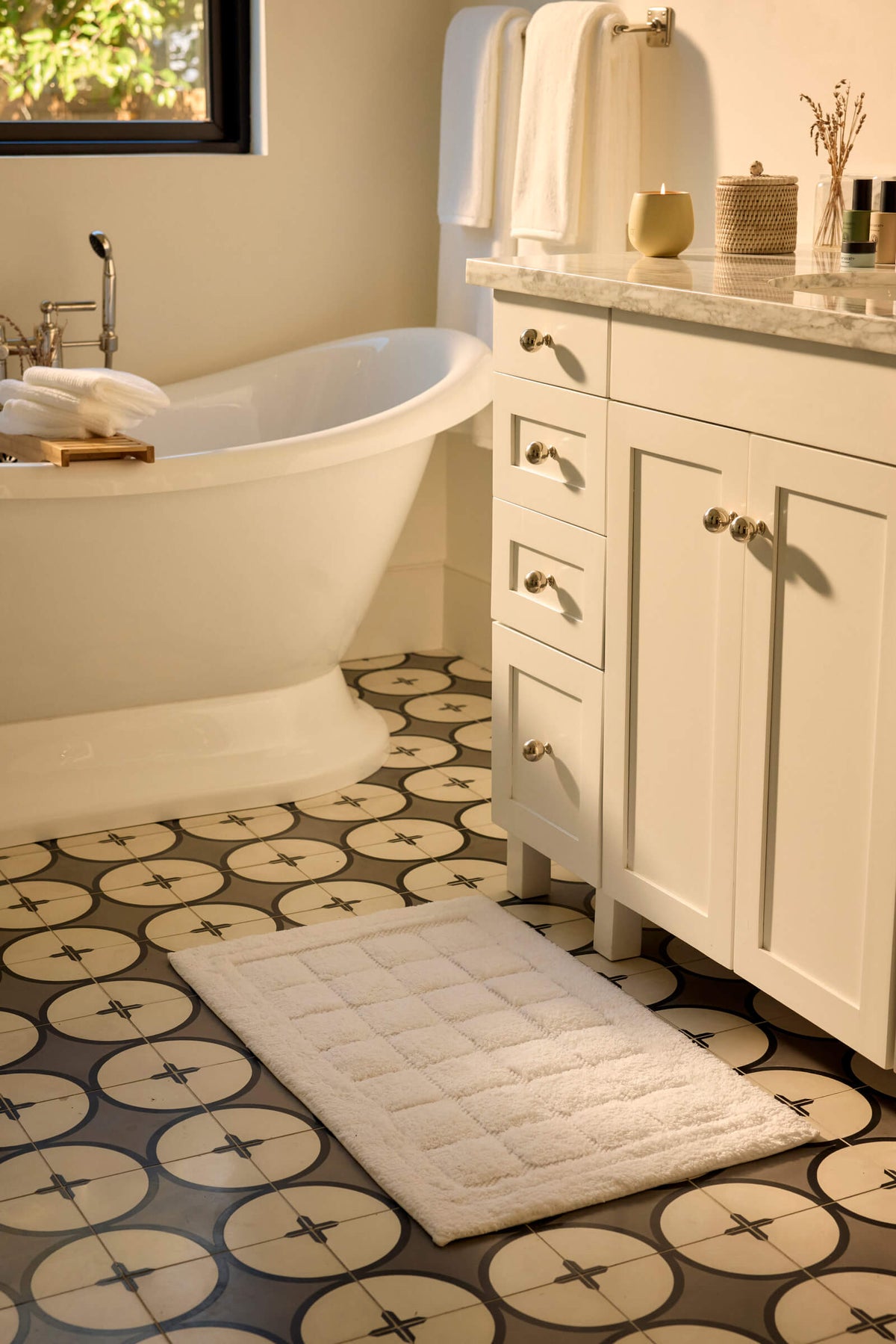 A bathroom with a patterned tile floor, a white freestanding bathtub, a white vanity with drawers, Cozy Earth's Grid Bath Mat, and neatly folded towels on the counter and towel bar. Sunlight filters in from a window.|Color:White