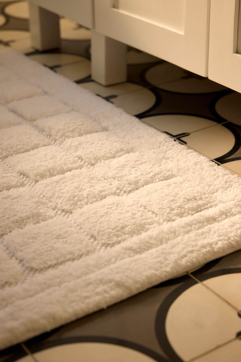 A close-up of the Cozy Earth Grid Bath Mat, featuring a white textured square pattern, placed on a circular black and white tiled floor next to a white cabinet.|Color:White