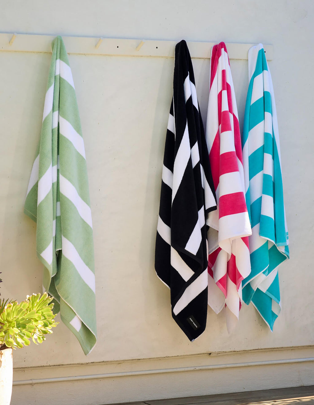 Four Cozy Earth Seaside Resort Towels in green, black, pink, and blue hang side by side on wall hooks. A potted succulent is visible in the lower left corner of the image.|Color:Aqua Stripe