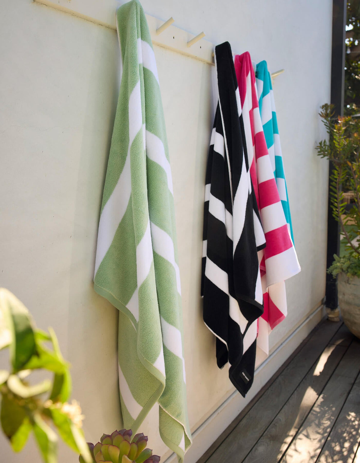 Four Cozy Earth Classic Stripe Resort Towels in green, black, red, and blue hang on hooks outside against a white wall, with plants and wooden decking nearby.|Color:Julep