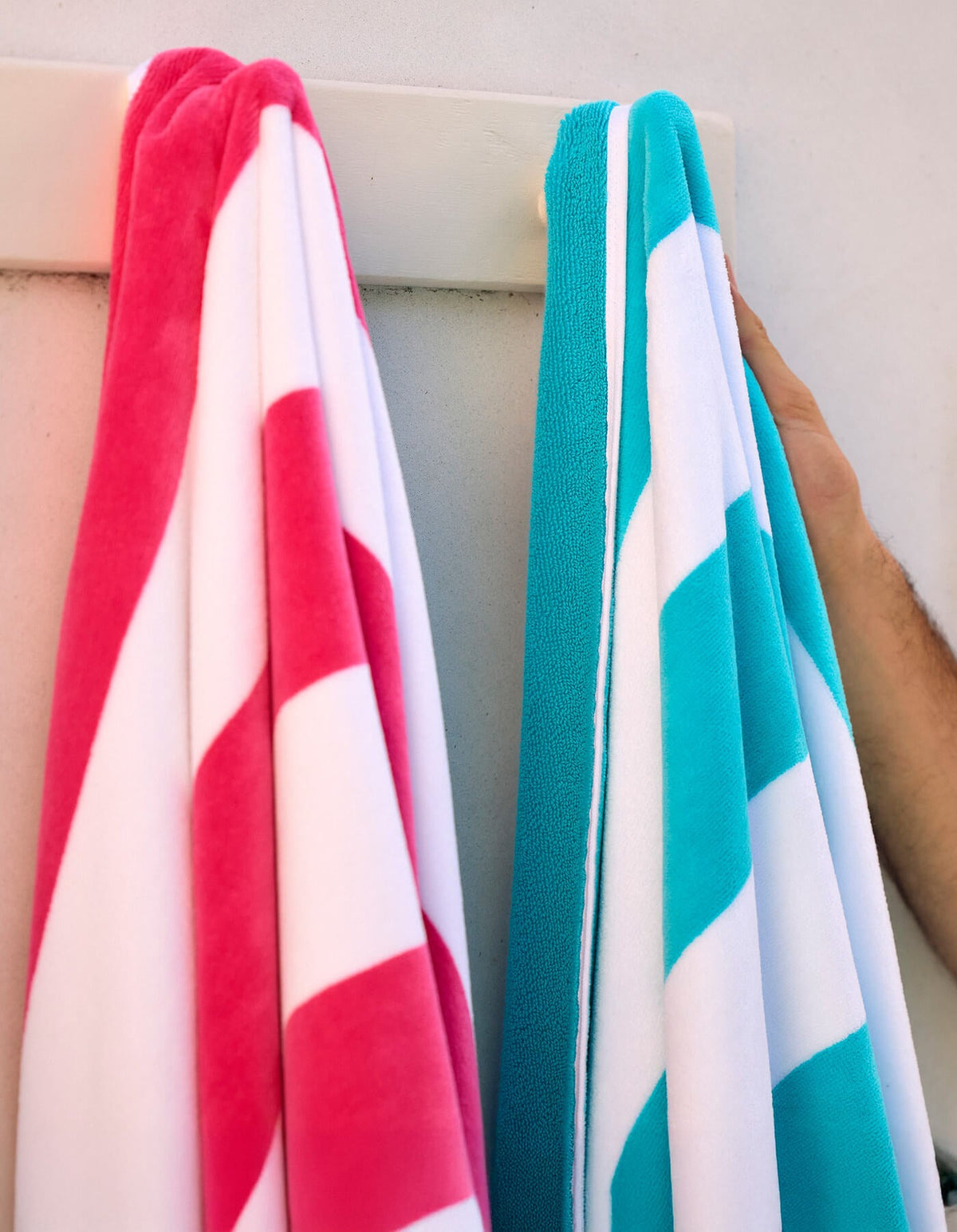 Two Cozy Earth Seaside Resort Towels—one pink and white, the other blue and white—hang on a white rack against a light wall. A hand holds the blue towel.|Color:Aqua Stripe