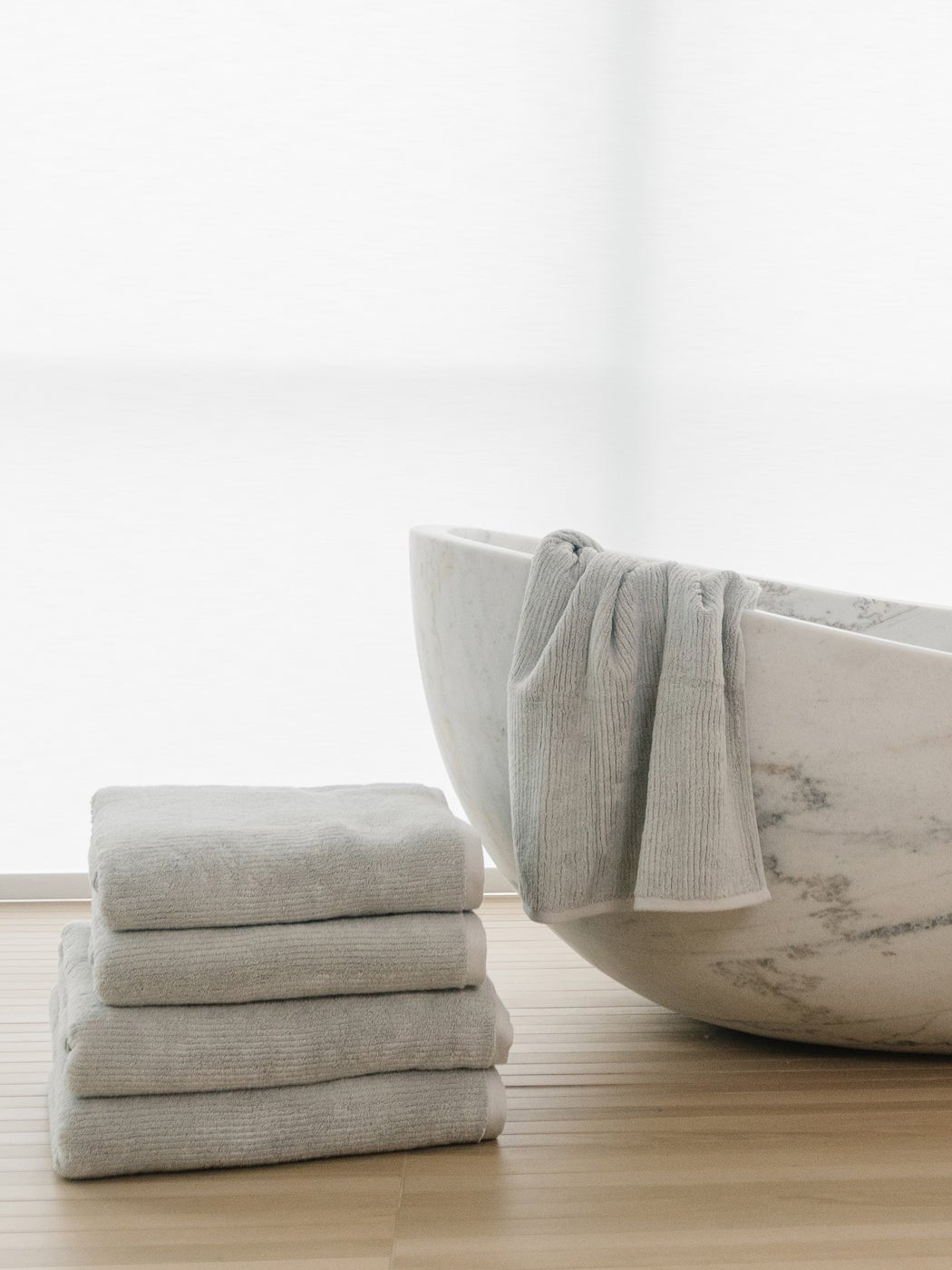 The Complete Ribbed Terry Bath Bundle by Cozy Earth features four neatly folded gray towels stacked next to a white marble bathtub, with another towel draped over the edge, all on a light wooden floor in a bright, minimalist setting.|Color:Light Grey