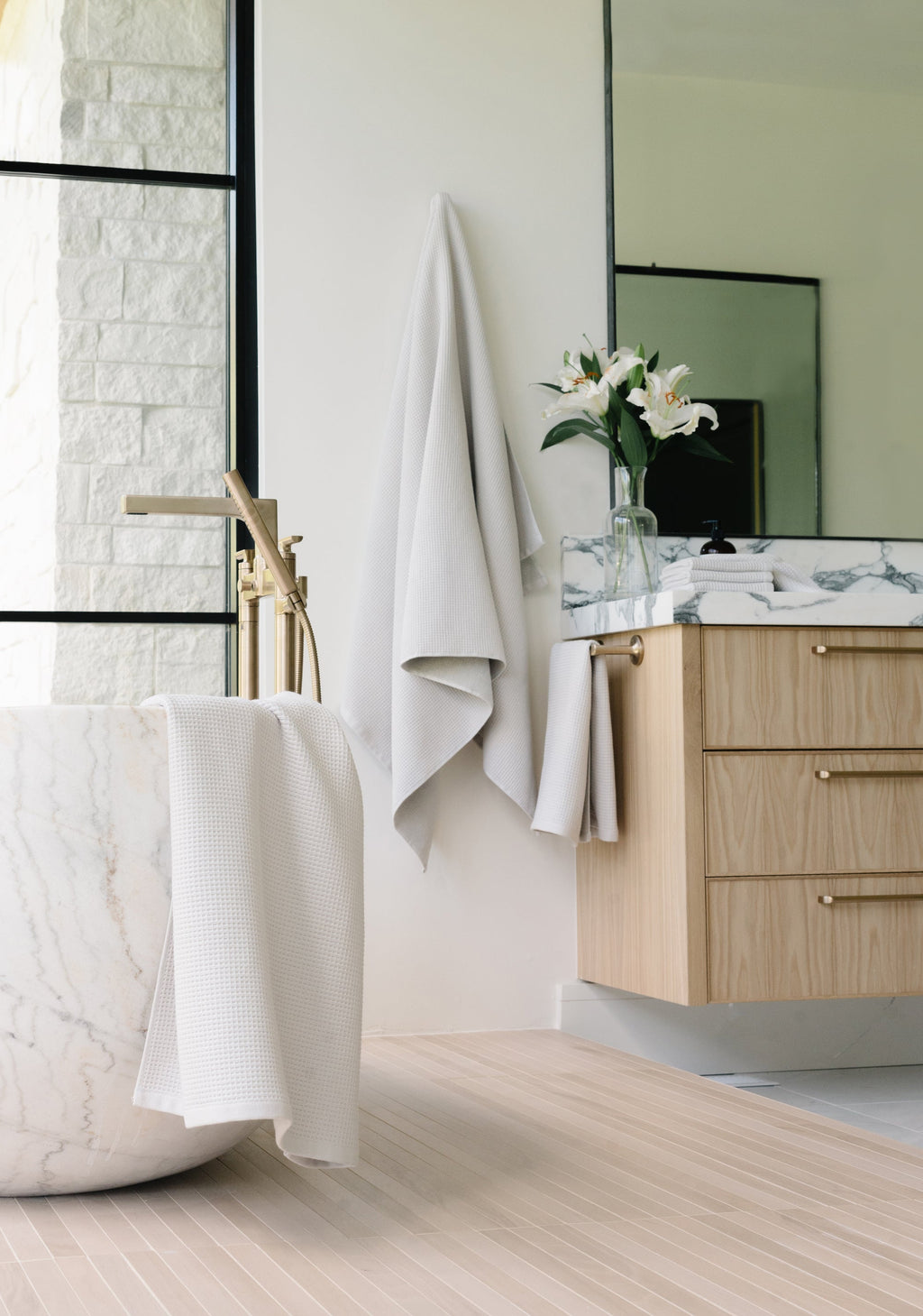 Light Grey Waffle Bath Sheet. The Waffle Bath Sheet in the color Light Grey is photographed in the bathroom. Photo of Light Grey Waffle Bath Towel taken hanging from a towel hook. The towel hook is not seen. A bath tub with another waffle towel draped over the tub is seen in the photo.|Color:Light Grey