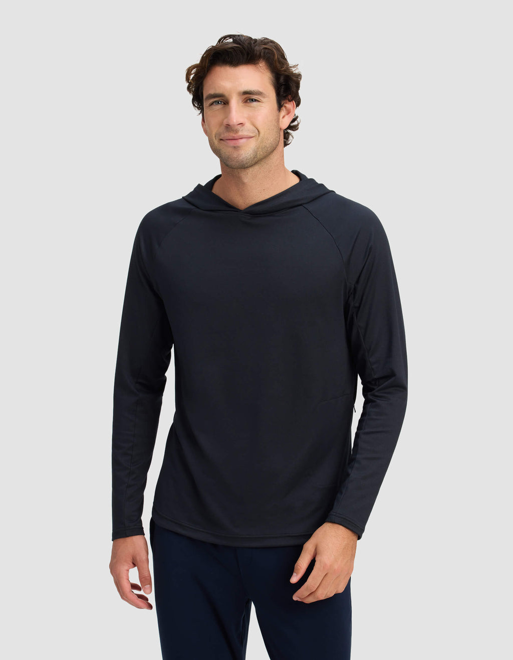 A man with short, wavy brown hair is wearing the Men's Lightweight Performance Hoodie by Cozy Earth, standing against a plain light gray background and smiling slightly at the camera.|Color:Jet Black