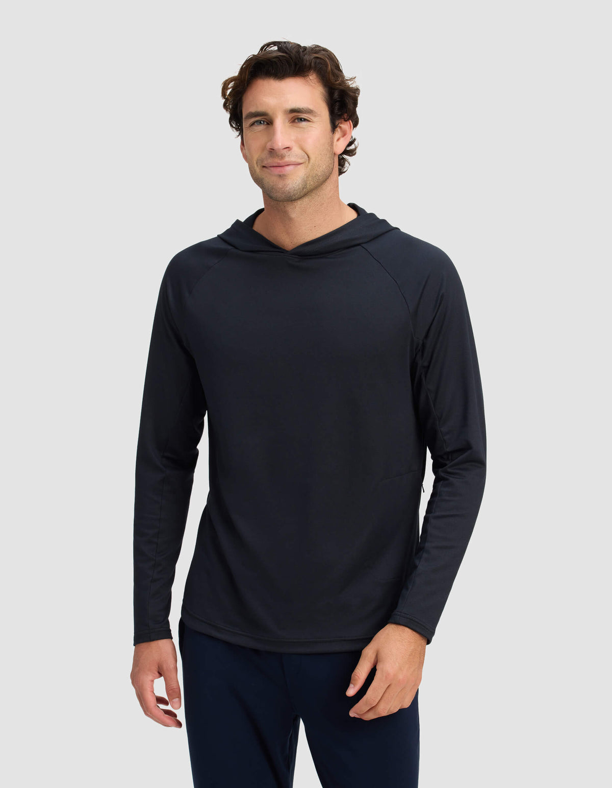 A man with short, wavy brown hair is wearing the Men's Lightweight Performance Hoodie by Cozy Earth, standing against a plain light gray background and smiling slightly at the camera.|Color:Jet Black