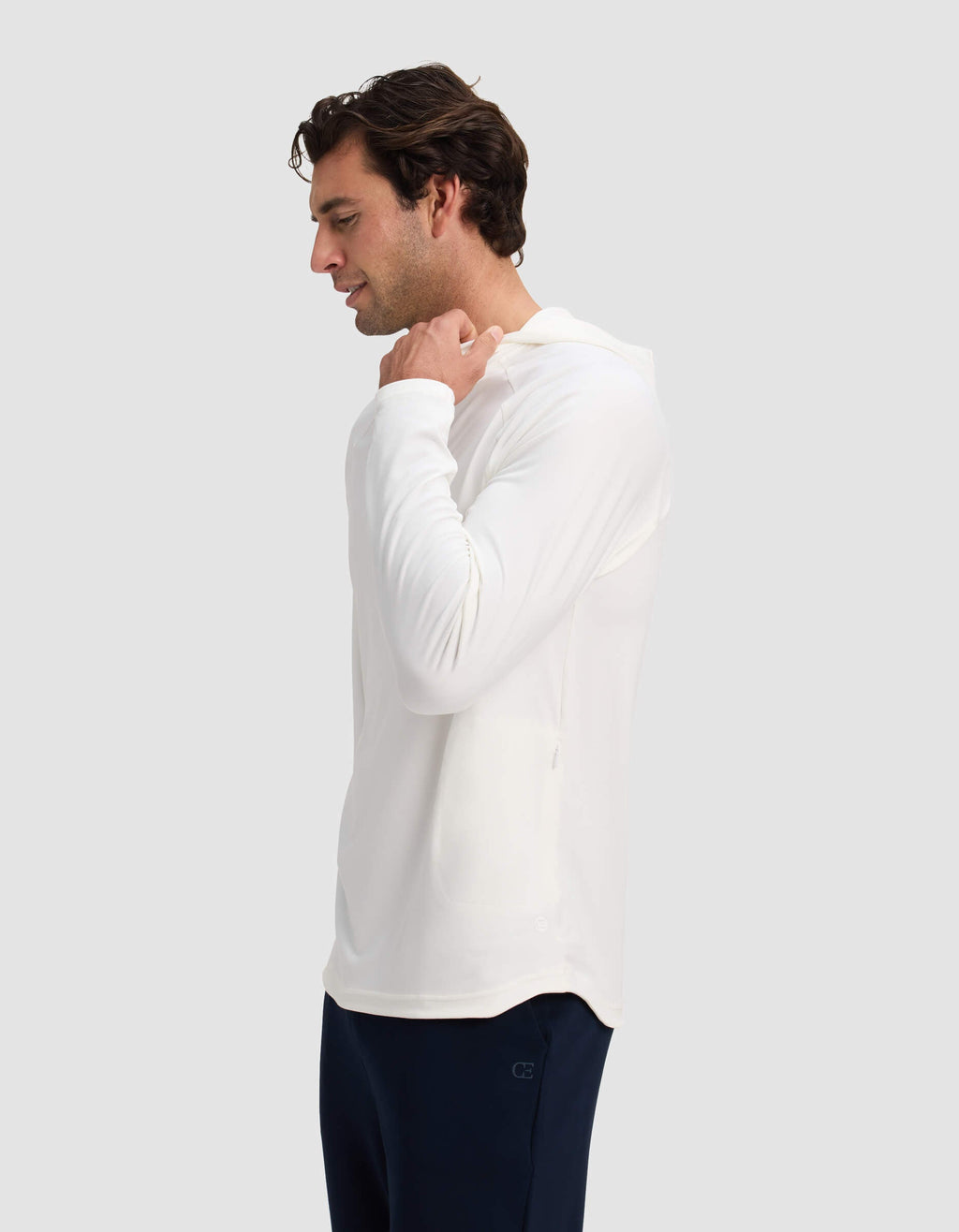A man wearing the Cozy Earth Men's Lightweight Performance Hoodie in white and dark pants faces left, holding the hood with one hand against a plain light gray background.|Color:White