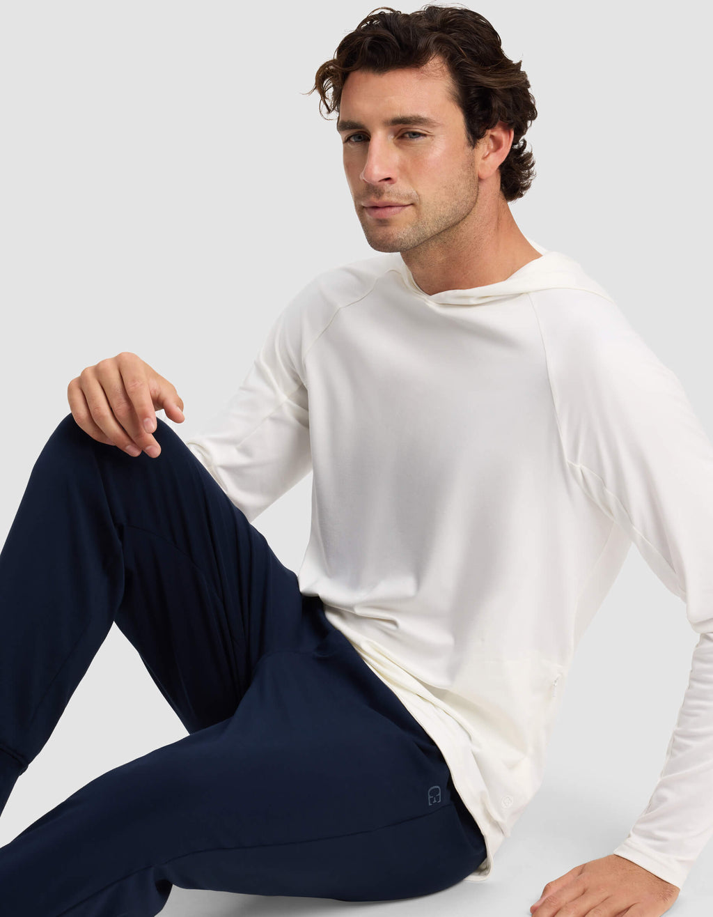 A man with wavy brown hair sits on the floor in a long-sleeve white Men's Lightweight Performance Hoodie by Cozy Earth and dark blue pants. He looks at the camera with a neutral expression, against a plain light gray background.|Color:White