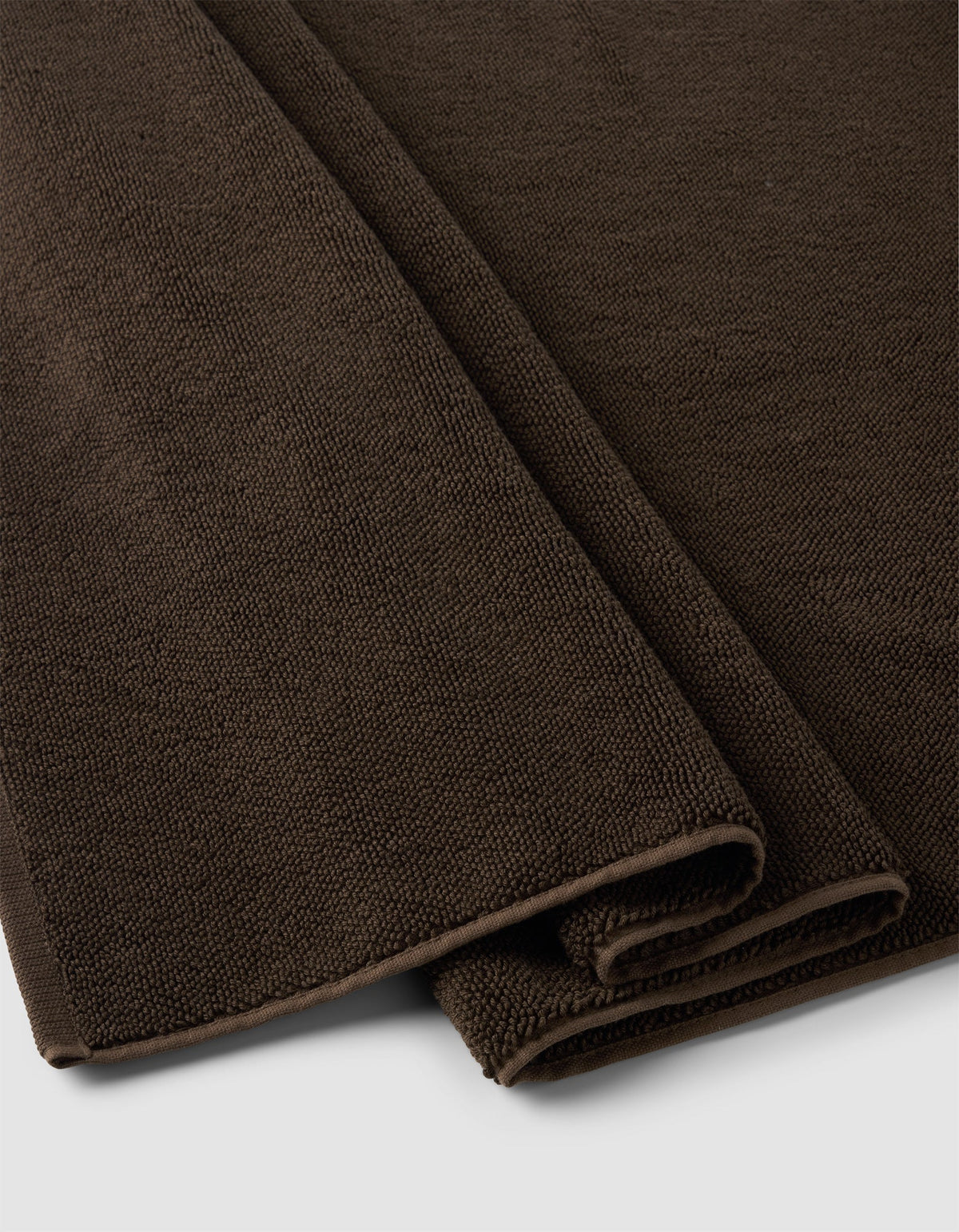 A close-up of two folded Cozy Earth Loop Terry Bath Mats in dark brown, featuring textured woven fabric, placed on a light gray surface.|Color:Espresso