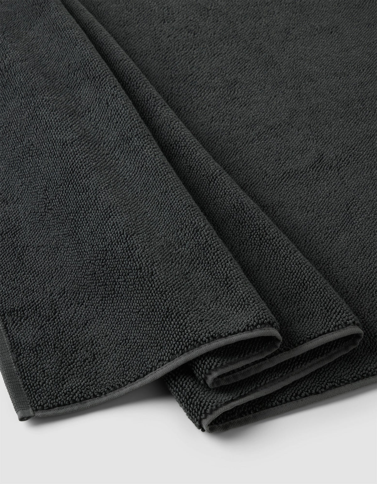 A close-up of the Cozy Earth Loop Terry Bath Mat, neatly folded in dark gray with a soft, textured surface, displayed on a light-colored background.|Color:Graphite
