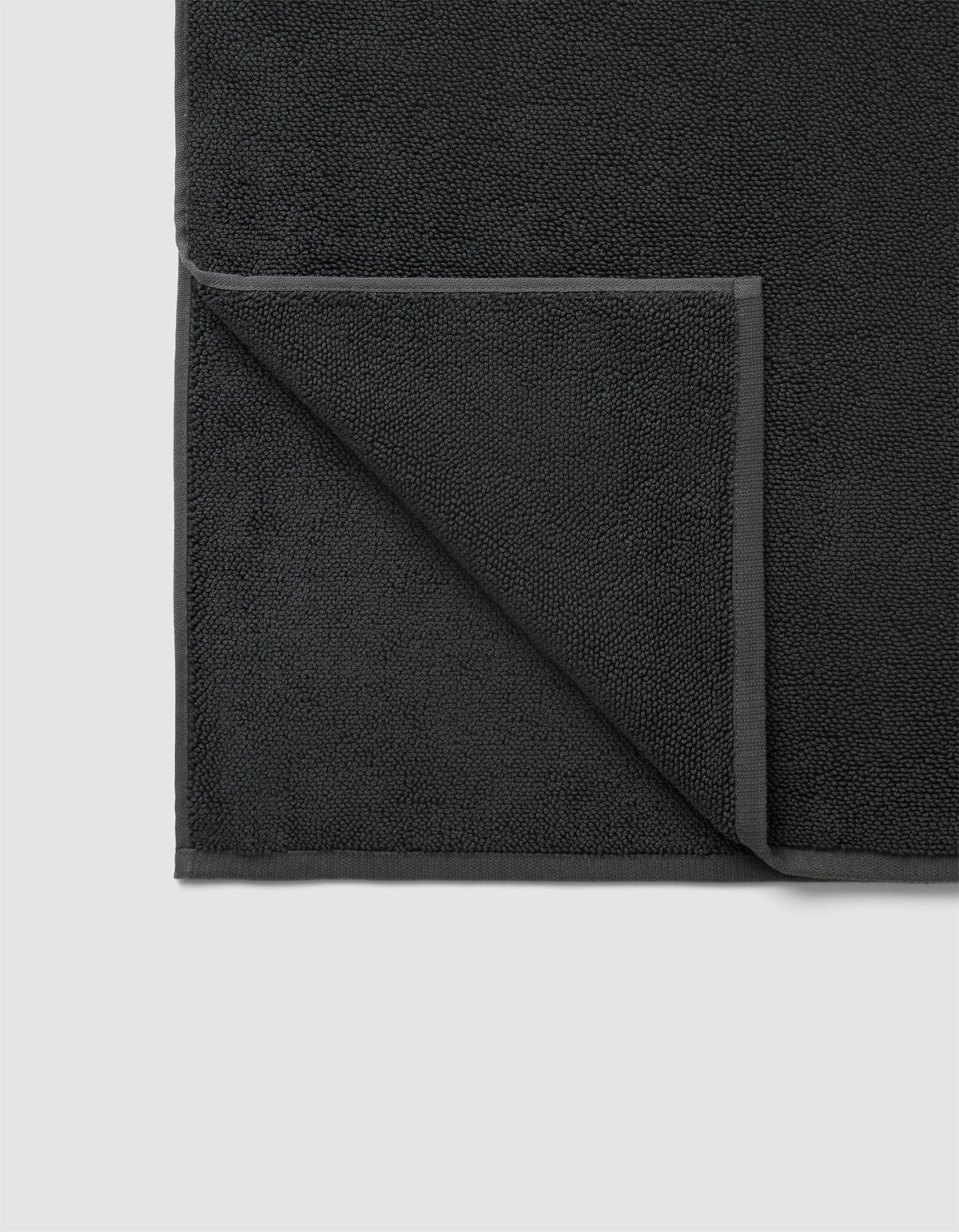 The Cozy Earth Loop Terry Bath Mat in dark gray is pictured neatly folded at one corner, displaying its textured surface and smooth edge border on both front and back sides.|Color:Graphite