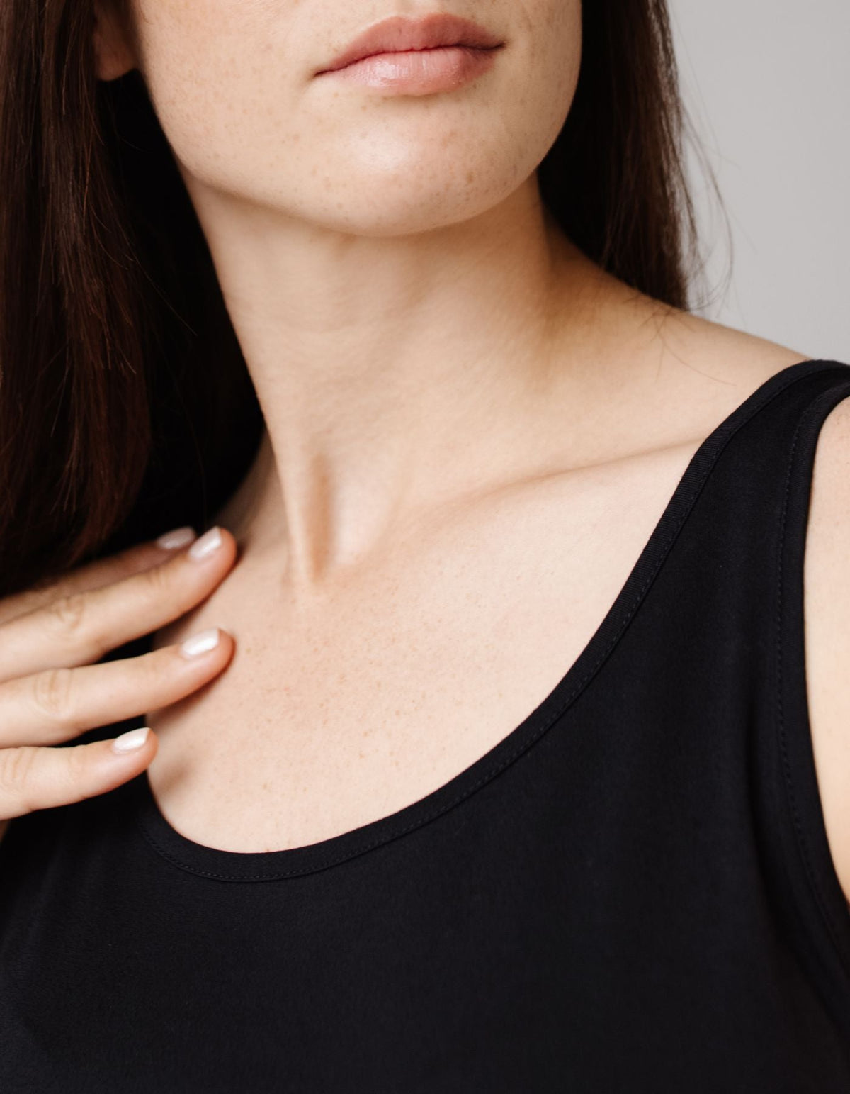A close-up shows a woman wearing the Cozy Earth Women's Bamboo Stretch-Knit Lounge Tank in black, touching her collarbone with her left hand. Only her lower face, neck, and upper chest are visible.
