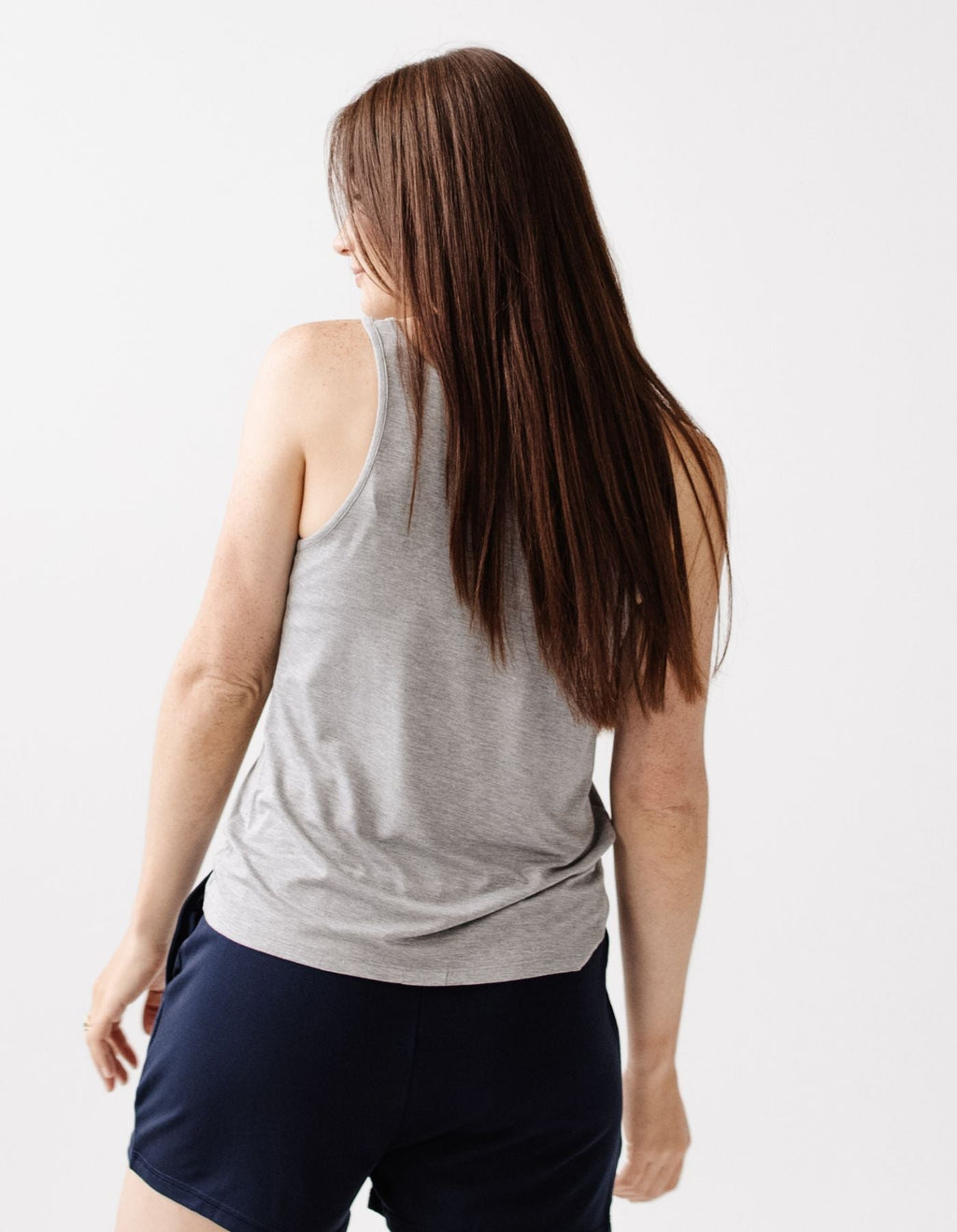 A woman with long brown hair wears the Cozy Earth Women's Bamboo Stretch-Knit Lounge Tank in gray, paired with navy shorts, standing against a plain white background and looking slightly over her shoulder.|Color:Grey