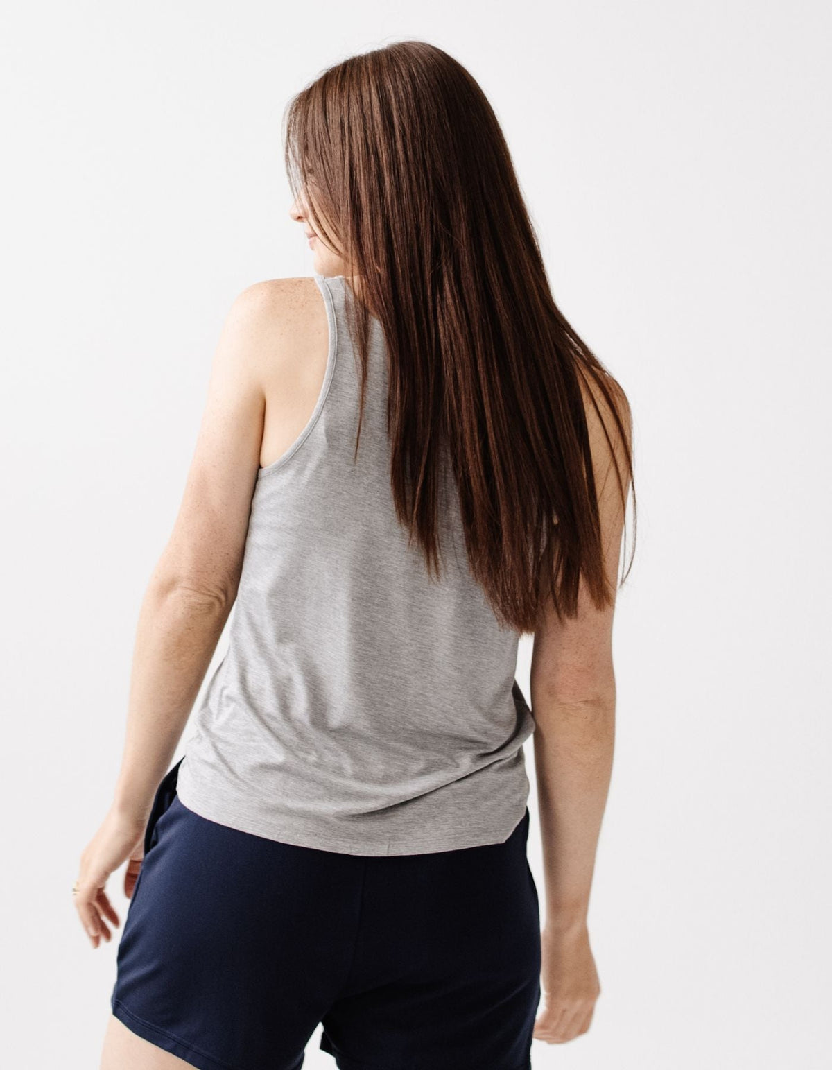 A woman with long brown hair wears the Cozy Earth Women's Bamboo Stretch-Knit Lounge Tank in gray, paired with navy shorts, standing against a plain white background and looking slightly over her shoulder.|Color:Grey
