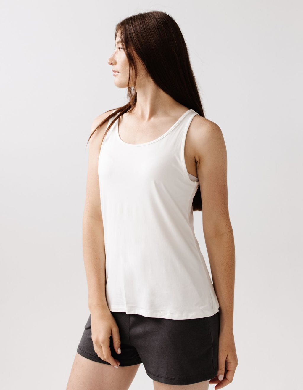 A woman with long brown hair wears the Cozy Earth Women's Bamboo Stretch-Knit Lounge Tank in Ivory, paired with dark shorts, standing against a plain light background and looking to her left.|Color:Ivory