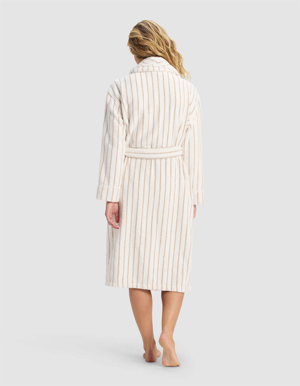 A woman with wavy blonde hair stands barefoot on a plain light background, shown from the back in Cozy Earth's Striped Luxe Bath Robe—a long, cream robe with beige vertical stripes and a belt.|Color:Sand Stripe