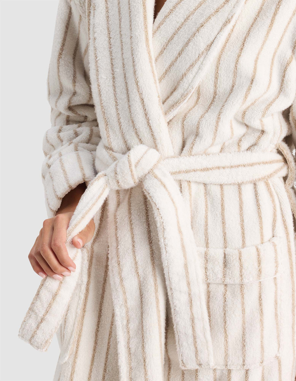 A person wears the Cozy Earth Striped Luxe Bath Robe, a soft cream-colored robe with beige vertical stripes, tied at the waist; only their upper body and one hand adjusting the belt are visible.|Color:Sand Stripe