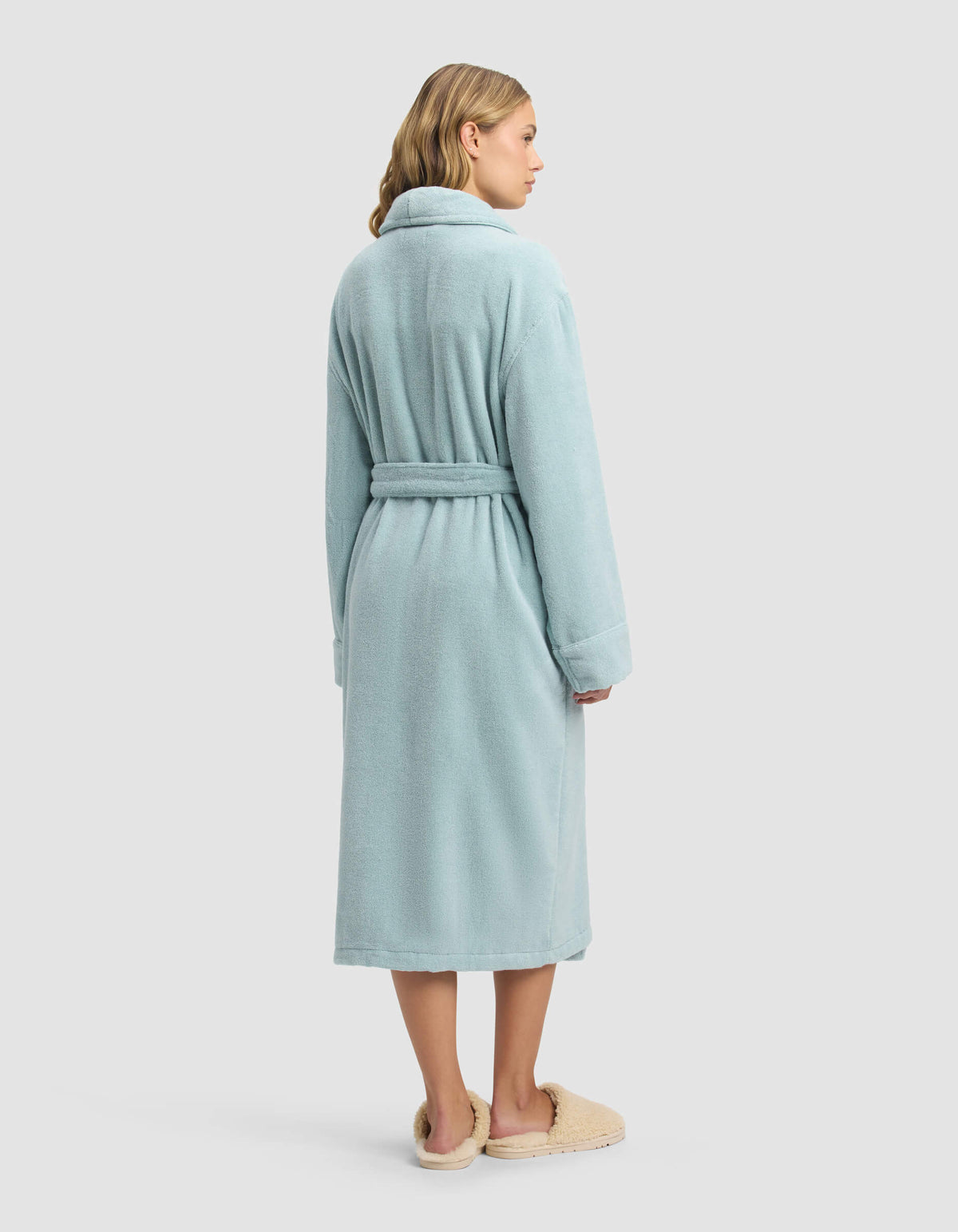 A woman with shoulder-length blonde hair stands facing away against a white background, wearing the Cozy Earth Luxe Bath Robe in light blue with a belt, paired with beige slippers.|Color:Tide