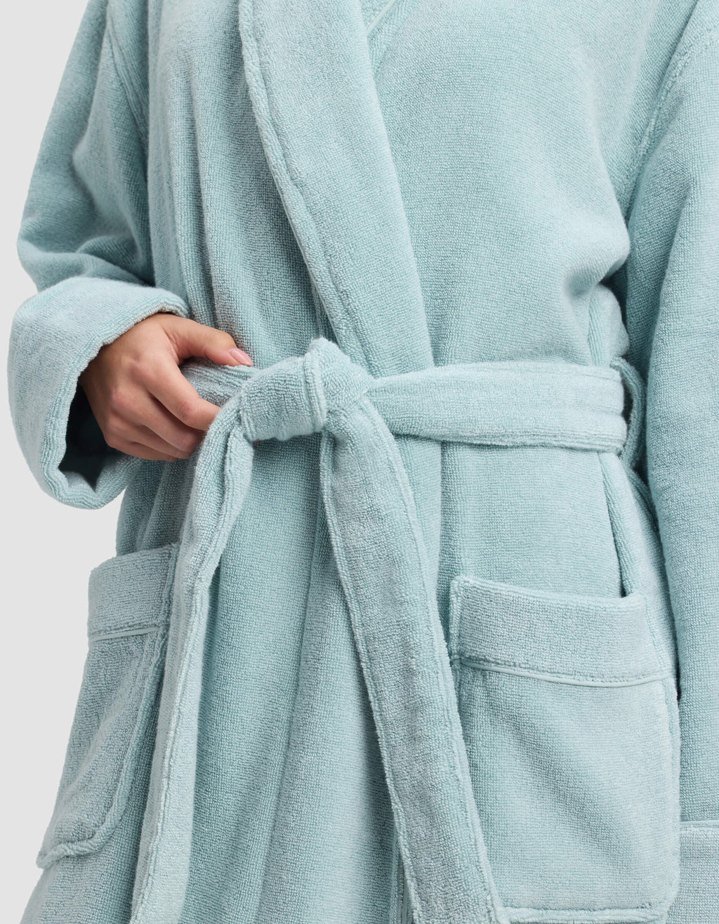 A person wearing the Cozy Earth Luxe Bath Robe in light blue ties its belt at the waist, showing only their torso and one hand, with two front pockets visible.|Color:Tide