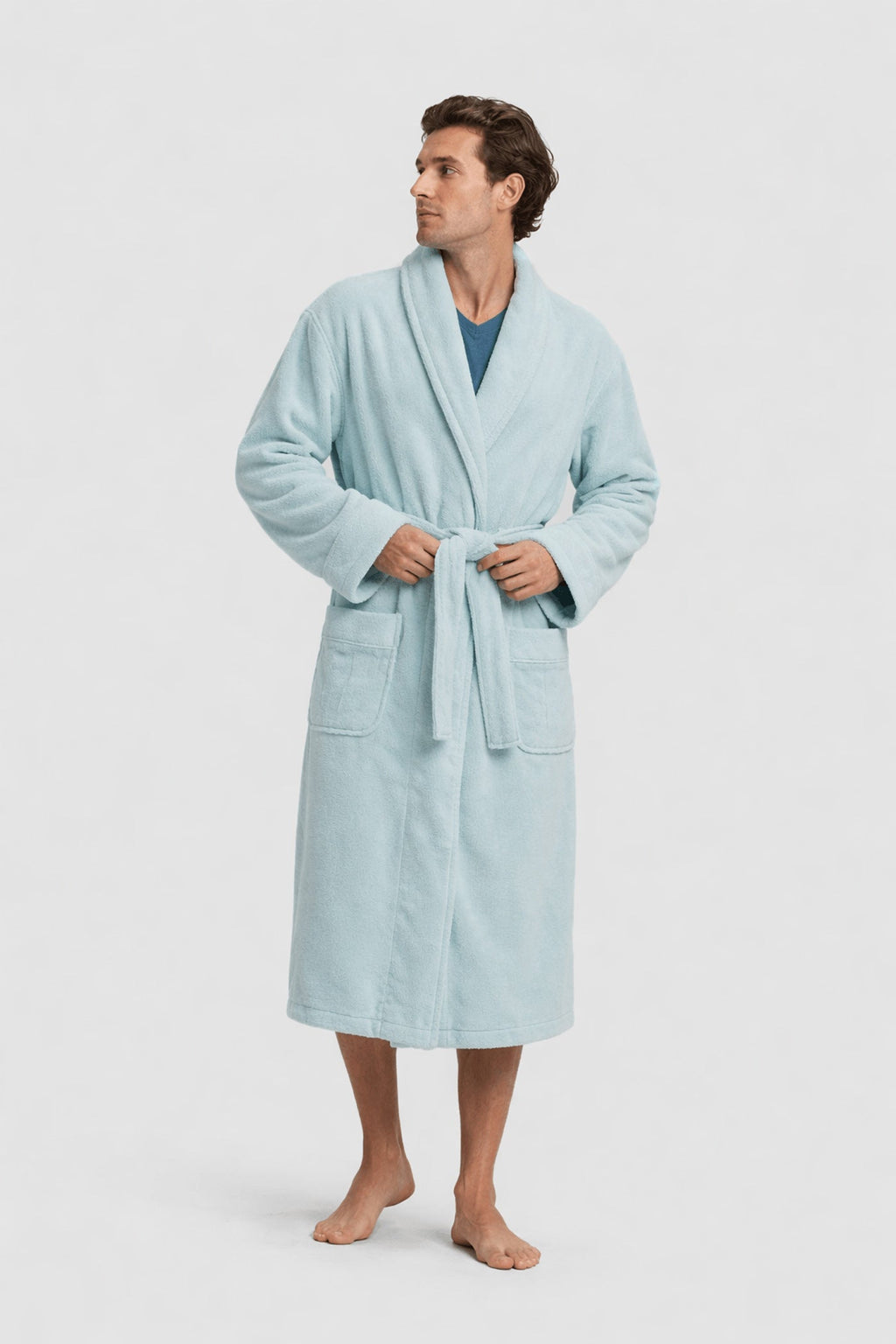 A man stands barefoot in the Luxe Bath Robe by Cozy Earth—a light blue, knee-length robe with a belt and two front pockets—over a blue shirt, looking to his left against a plain light background.|Color:Tide