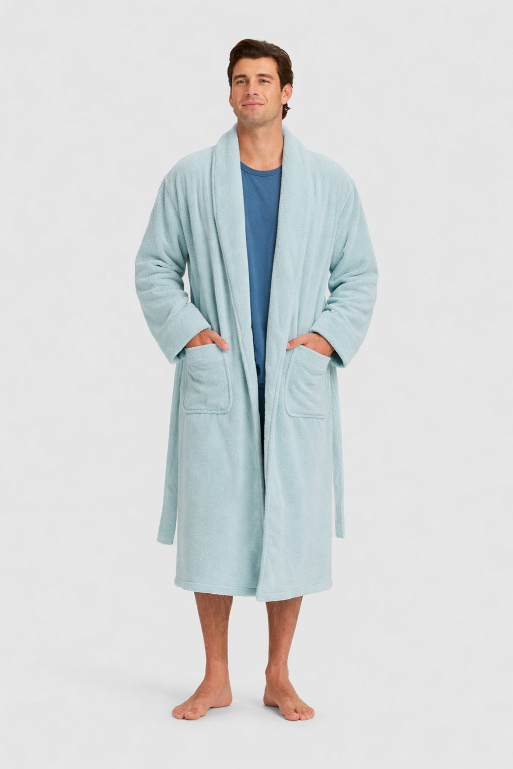 A man stands barefoot in a Luxe Bath Robe by Cozy Earth over a blue shirt, hands in the robe’s pockets, smiling gently against a plain light background.|Color:Tide