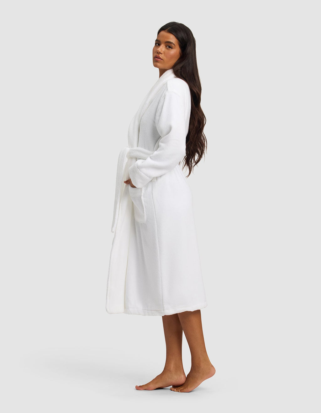 A woman with long dark hair stands barefoot on a white background, wearing the Luxe Bath Robe by Cozy Earth, tied at the waist. She faces slightly to the side with her hands in the robe’s pockets.|Color:White