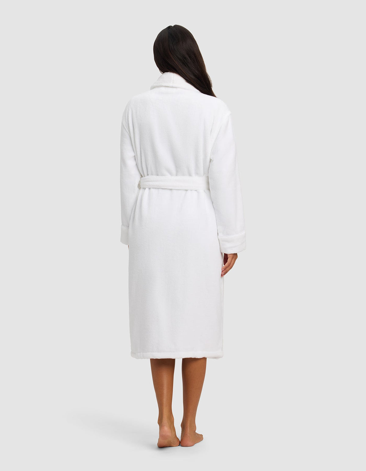 A woman with long dark hair stands barefoot facing away, wearing the Cozy Earth Luxe Bath Robe—knee-length, white, and belted—against a plain light background.|Color:White