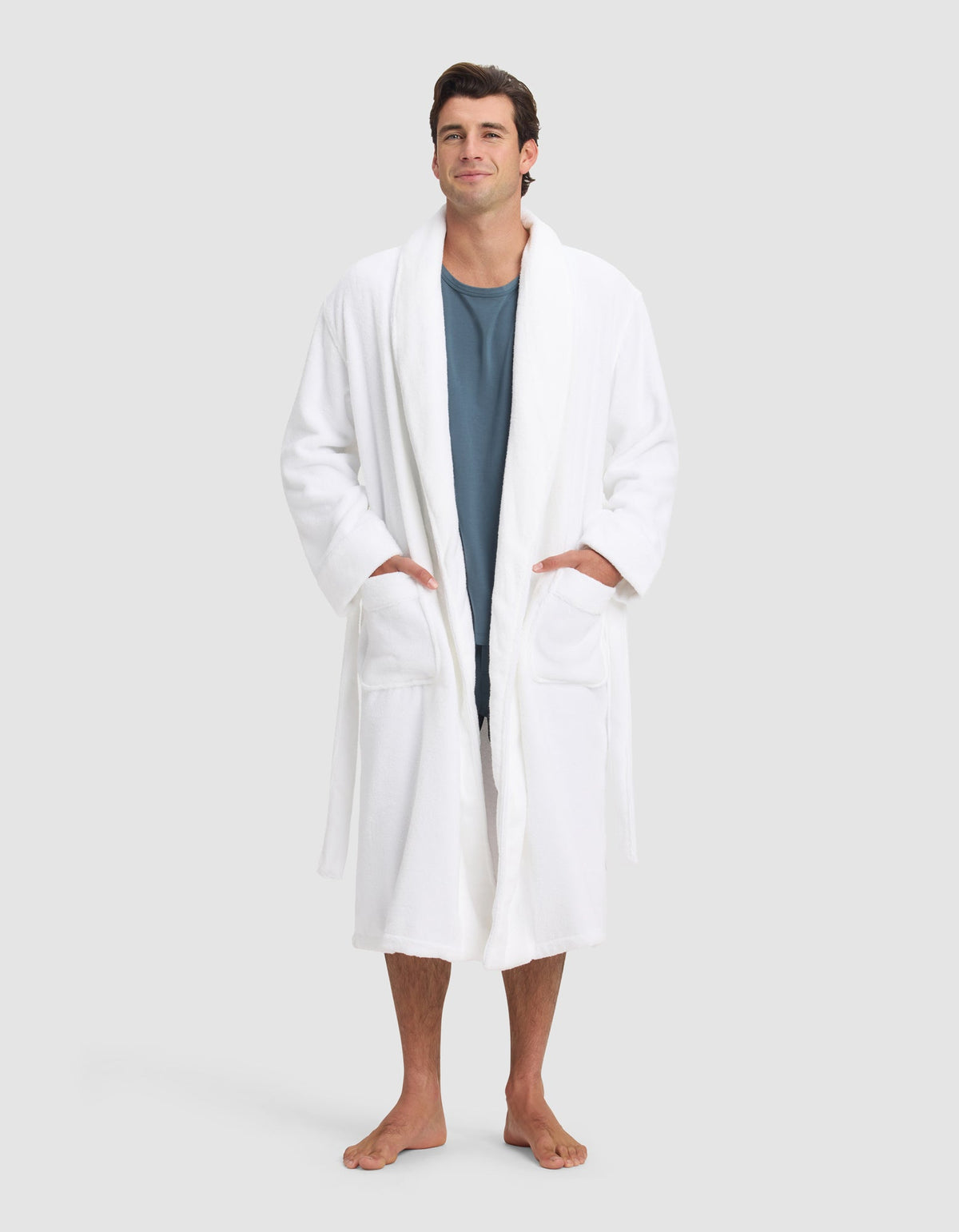 A man stands barefoot against a neutral background, wearing the Cozy Earth Luxe Bath Robe over a blue shirt. His hands are in the robe’s pockets as he looks forward with a slight smile.|Color:White