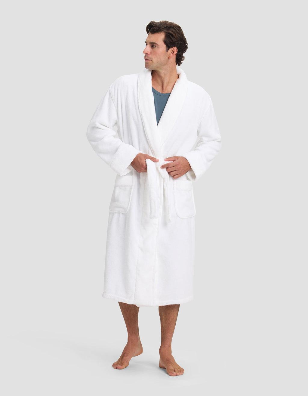 A man with short, dark hair stands barefoot in a Cozy Earth Luxe Bath Robe over a blue shirt, tying the belt at his waist. He faces slightly right against a plain light gray background.|Color:White
